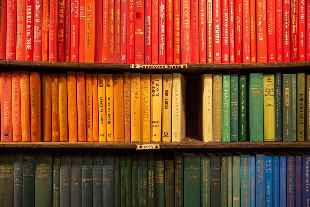 Three shelves of a library, showing books arranged by color of the spine. The top shelf is red books, the middle shelf is orange to green, the bottom shelf is dark green.